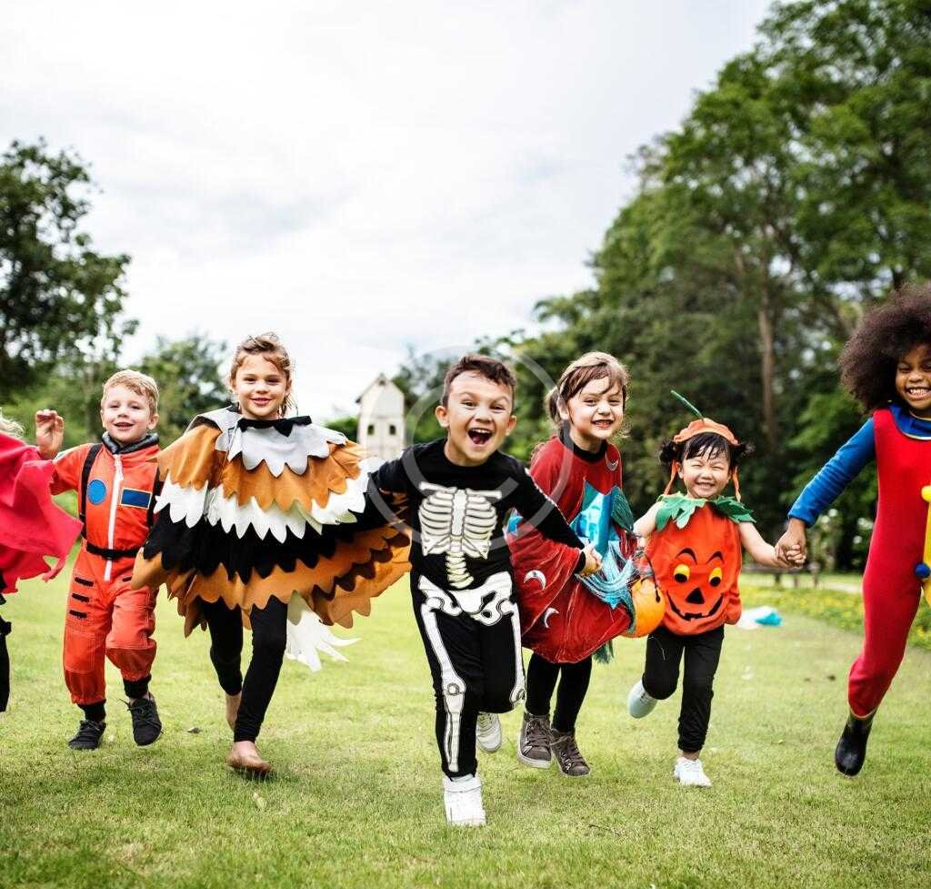 Children dressed in various costumes running and holding hands on a grassy field.