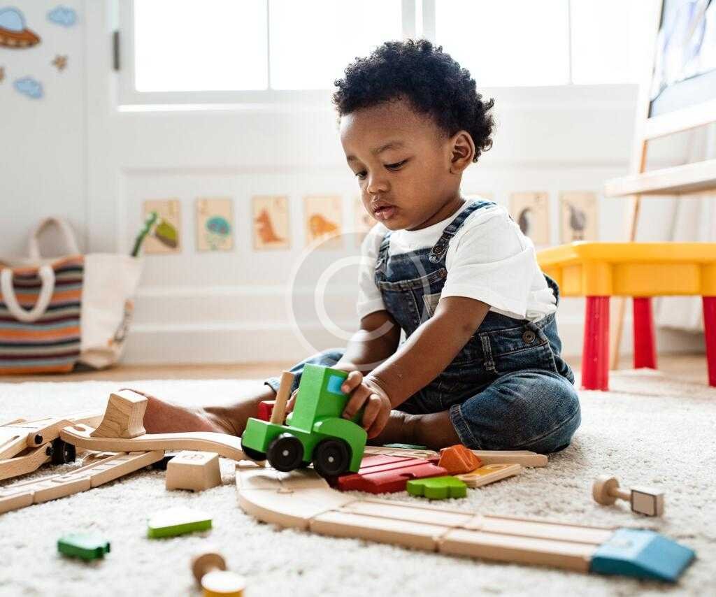 A child playing with wooden train toys on a carpeted floor in a brightly lit room.