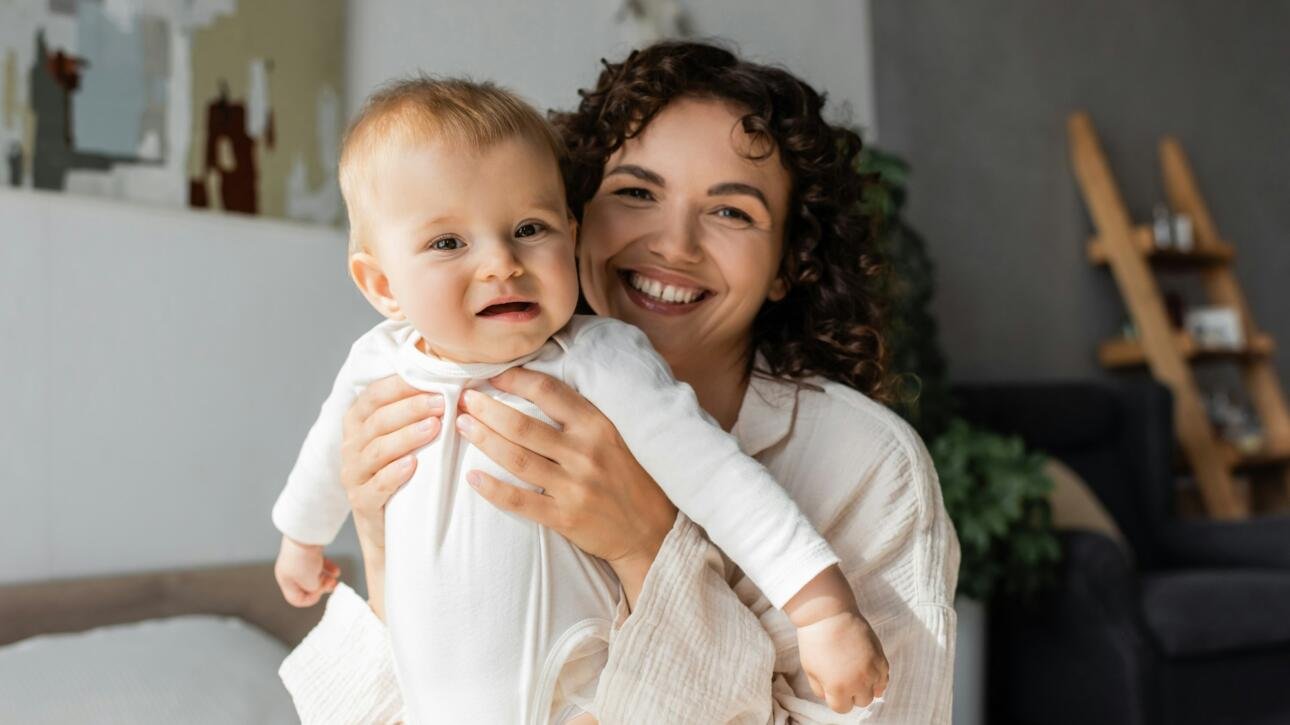 cheerful and curly mother holding daughter in romper in bedroom