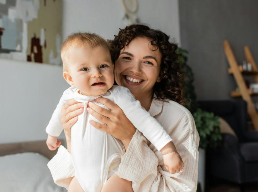 cheerful and curly mother holding daughter in romper in bedroom