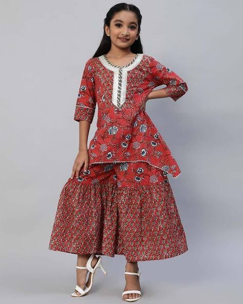 Young girl wearing a red floral printed kurta with matching tiered skirt and white sandals.