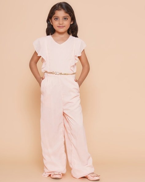 Girl wearing a light pink jumpsuit with a belt, standing with hands in pockets, against a beige background.