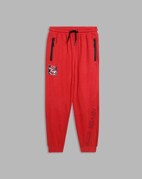 Red joggers with black drawstring, zippered pockets, patch detail, and "Never Stop" text.