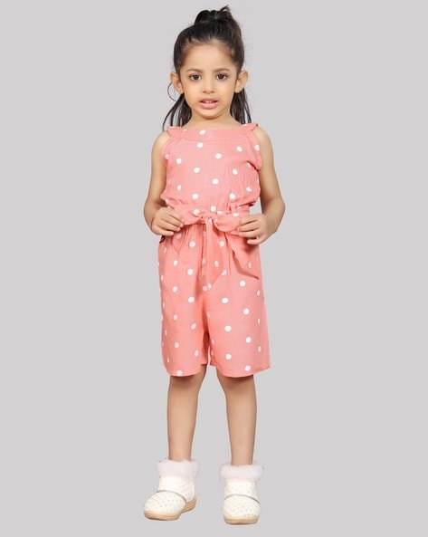 Young girl wearing a pink polka dot romper with white shoes against a grey background.