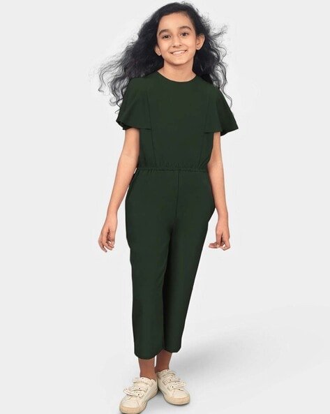 Young girl in a dark green jumpsuit and beige trainers, standing and smiling.