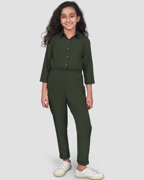 A girl in a green button-up jumpsuit and white sneakers, standing and smiling.