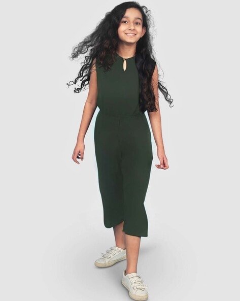 A person with long dark hair wearing a sleeveless dark green dress and white sneakers is smiling and walking forward.